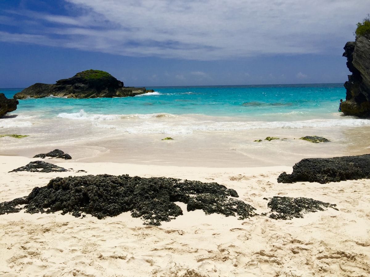 Turquoise cove with black limestone rock formations and a small islet off a white sand Bermuda beach