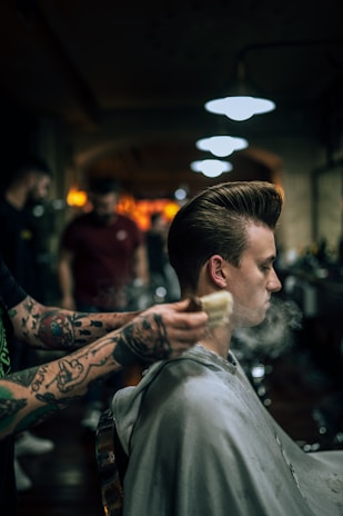 A person is sitting in a barber chair getting a haircut. The barber's tattooed arms are visible as they brush the client's hair. The setting is a dimly lit barbershop with several people and warm lighting in the background. Steam or powder is visible around the client's head, suggesting grooming products are being used.