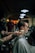 A person is sitting in a barber chair getting a haircut. The barber's tattooed arms are visible as they brush the client's hair. The setting is a dimly lit barbershop with several people and warm lighting in the background. Steam or powder is visible around the client's head, suggesting grooming products are being used.