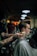 A person is sitting in a barber chair getting a haircut. The barber's tattooed arms are visible as they brush the client's hair. The setting is a dimly lit barbershop with several people and warm lighting in the background. Steam or powder is visible around the client's head, suggesting grooming products are being used.