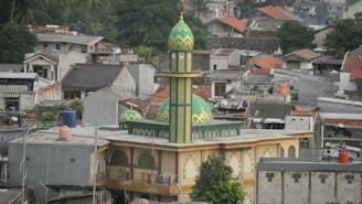 A green and yellow mosque with a tall minaret is situated in the middle of an urban residential area. Surrounding the mosque are numerous houses and buildings with various architectural styles, some with red roofs and some with flat roofs. There is greenery, including trees, visible around the houses.