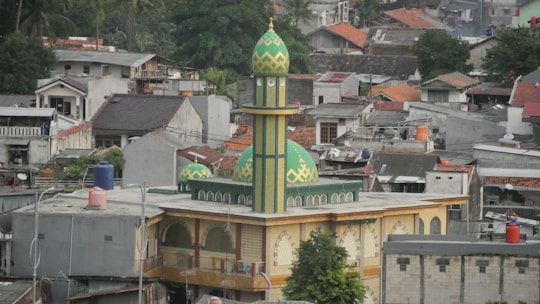 A green and yellow mosque with a tall minaret is situated in the middle of an urban residential area. Surrounding the mosque are numerous houses and buildings with various architectural styles, some with red roofs and some with flat roofs. There is greenery, including trees, visible around the houses.
