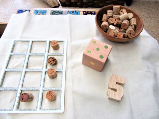A wooden tic-tac-toe game board with cork pieces is placed on a light fabric. A wooden bowl filled with additional corks is nearby, along with a large wooden die featuring green dots and a wooden number 5.