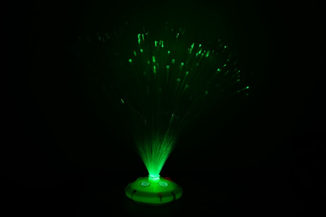 A fiber optic lamp emits an array of glowing green lights. The slender fibers fan out from the base, creating an enchanting display of shimmering green points that contrast with the darkness around.