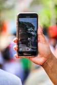 A hand is holding a smartphone with a video recording interface on display. The screen depicts an outdoor scene with people and trees, suggesting an event or gathering. The background is blurred, emphasizing focus on the phone and its content.
