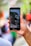 A hand is holding a smartphone with a video recording interface on display. The screen depicts an outdoor scene with people and trees, suggesting an event or gathering. The background is blurred, emphasizing focus on the phone and its content.