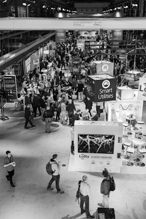 A bustling trade show or conference with numerous attendees interacting around various exhibition booths. The venue is spacious with high ceilings and multiple displays, including one prominently featuring Smart Utilities and another labeled with LoRa technology. People are engaged in discussions and networking, adding to the lively atmosphere.