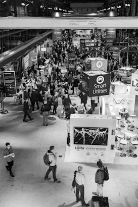A bustling trade show or conference with numerous attendees interacting around various exhibition booths. The venue is spacious with high ceilings and multiple displays, including one prominently featuring Smart Utilities and another labeled with LoRa technology. People are engaged in discussions and networking, adding to the lively atmosphere.