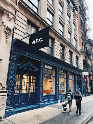 A street scene featuring a stylish storefront with a navy blue facade and large windows displaying interior items. A black flag with 'A.P.C.' is prominently displayed above the entrance. Two people walk by with a dog on the sidewalk in front of the store, and neighboring buildings are visible with fire escape staircases.
