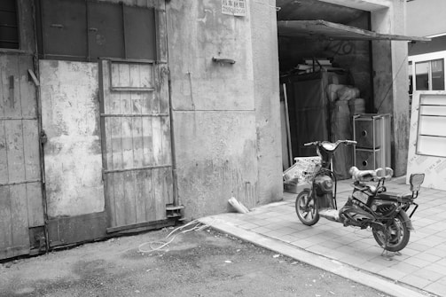 A black and white photograph depicts an electric scooter parked in front of a weathered, industrial building. The building features a set of large, metal, and wooden doors with visible wear and rust. Inside, the building appears cluttered, with stacked items and furniture partly visible through the open entrance.