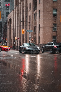 A wet urban street scene with cars driving through falling snow. The buildings are tall and modern, with windows and signs visible. A Canadian flag and traffic lights add to the bustling city atmosphere, while reflections on the wet road create an almost cinematic effect.