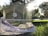 A birdcage sits on the grass in a sunlit garden. The sun is shining brightly in the background, casting a warm glow over the scene. Trees and a path are visible in the background. A parakeet is perched inside the cage, which is partially covered with a purple cloth.