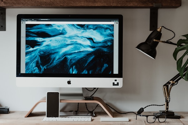 A modern desk setup featuring an Apple iMac displaying a vivid blue abstract artwork on the screen. The desk includes a smartphone docked in front of the monitor, a sleek black desk lamp, a keyboard, glasses, and leafy green plant, creating a harmonious and organized workspace.