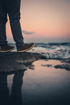 person in blue denim jeans and brown shoes standing on rocky shore during daytime