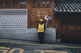 A person stands on a sidewalk in front of a traditional building with intricate wooden doors and tiled roof. They are wearing a yellow vest and hat, holding a sign that requests silence, emphasizing respect in a cultural setting.