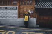A person stands on a sidewalk in front of a traditional building with intricate wooden doors and tiled roof. They are wearing a yellow vest and hat, holding a sign that requests silence, emphasizing respect in a cultural setting.