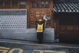 A person stands on a sidewalk in front of a traditional building with intricate wooden doors and tiled roof. They are wearing a yellow vest and hat, holding a sign that requests silence, emphasizing respect in a cultural setting.