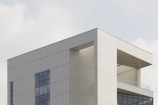 A modern building with a minimalist design characterized by sharp angles and a predominantly white facade. Large windows punctuate the smooth surface, allowing light to enter the interior spaces. The architectural style emphasizes open, unobstructed lines and the use of geometric shapes.