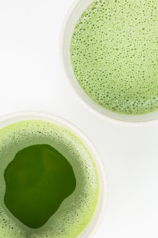 Two cups are filled with green frothy liquid, likely matcha, on a white surface. The texture appears creamy with a layer of bubbles on top.