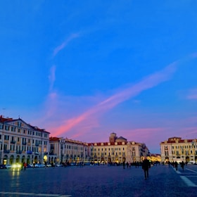 Evening view of a lively plaza surrounded by shops and apartments, illuminated by warm lights and active with residents.