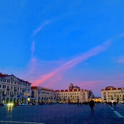 A large open plaza with classic multi-story buildings featuring arched windows and red-tiled roofs. The sky above is a vibrant mix of blue and pink, indicating a sunset. People are walking around the plaza, capturing a lively urban atmosphere.