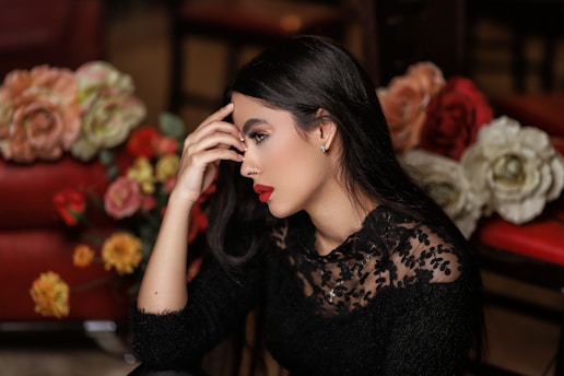 A woman with long dark hair and striking red lipstick is seated amidst a colorful arrangement of flowers. She appears thoughtful, with her hand gently resting on her temple. She is wearing a black lace top that adds an elegant touch to her appearance. The background is soft-focused, bringing the attention to the subject and the vibrant flowers surrounding her.