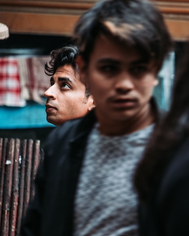 Two young men are in an urban setting with a focus on one man looking upward thoughtfully. The second man is slightly blurred in the foreground, adding a dynamic and introspective vibe.