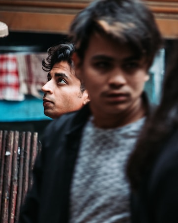 Two young men are in an urban setting with a focus on one man looking upward thoughtfully. The second man is slightly blurred in the foreground, adding a dynamic and introspective vibe.