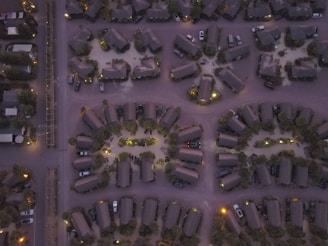An aerial view of a residential area with neatly arranged cabins and parked cars. The arrangement features a circular pattern, with the structures illuminated by soft outdoor lighting. The image is taken during dusk or dawn, casting a muted color palette over the scene.