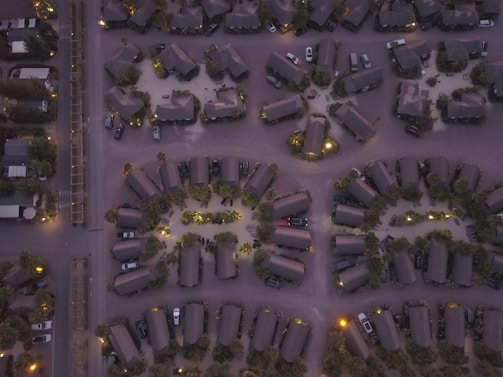 An aerial view of a residential area with neatly arranged cabins and parked cars. The arrangement features a circular pattern, with the structures illuminated by soft outdoor lighting. The image is taken during dusk or dawn, casting a muted color palette over the scene.