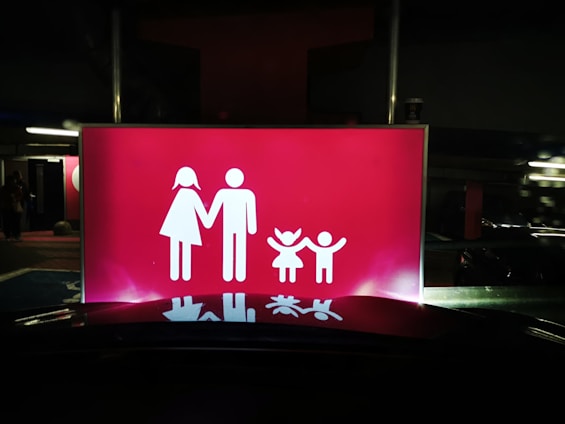 A bright red sign features white pictograms representing a family, consisting of two adults on the left and two children on the right, all holding hands. The scene is set against a dark background, suggesting it might be located in a parking area or similar environment.