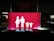 A bright red sign features white pictograms representing a family, consisting of two adults on the left and two children on the right, all holding hands. The scene is set against a dark background, suggesting it might be located in a parking area or similar environment.