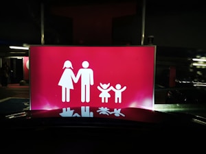 A bright red sign features white pictograms representing a family, consisting of two adults on the left and two children on the right, all holding hands. The scene is set against a dark background, suggesting it might be located in a parking area or similar environment.