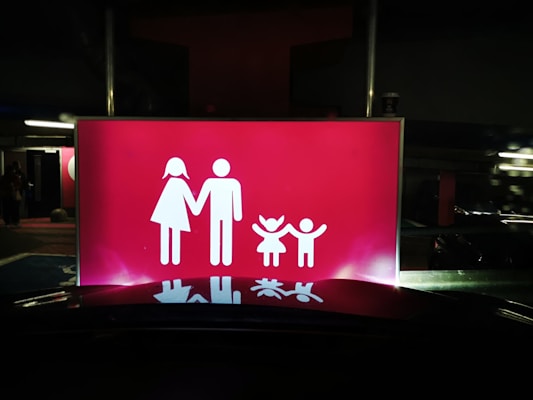 A bright red sign features white pictograms representing a family, consisting of two adults on the left and two children on the right, all holding hands. The scene is set against a dark background, suggesting it might be located in a parking area or similar environment.