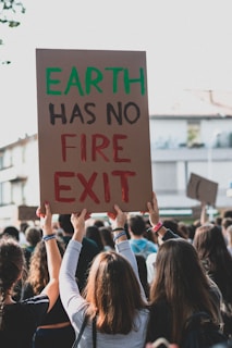 The image depicts a group of people participating in a protest. One of the protesters is holding up a cardboard sign that reads 'EARTH HAS NO FIRE EXIT' with the words 'EARTH' in green and 'FIRE EXIT' in red. The crowd is shown from the back, with the focus on the sign and the people raising their hands. The background shows some buildings and appears to be a street setting.