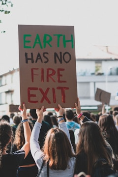 The image depicts a group of people participating in a protest. One of the protesters is holding up a cardboard sign that reads 'EARTH HAS NO FIRE EXIT' with the words 'EARTH' in green and 'FIRE EXIT' in red. The crowd is shown from the back, with the focus on the sign and the people raising their hands. The background shows some buildings and appears to be a street setting.