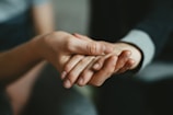 Two hands gently holding each other, conveying a sense of support and comfort. The background is blurred, focusing attention on the interaction between the hands.
