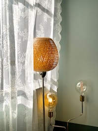 A woven lampshade emits a warm glow, standing beside a sheer lace curtain that features floral patterns. Additional exposed light bulbs add a modern touch with their visible filaments, while soft daylight filters through the lace, creating gentle shadows.