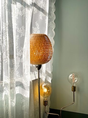A woven lampshade emits a warm glow, standing beside a sheer lace curtain that features floral patterns. Additional exposed light bulbs add a modern touch with their visible filaments, while soft daylight filters through the lace, creating gentle shadows.