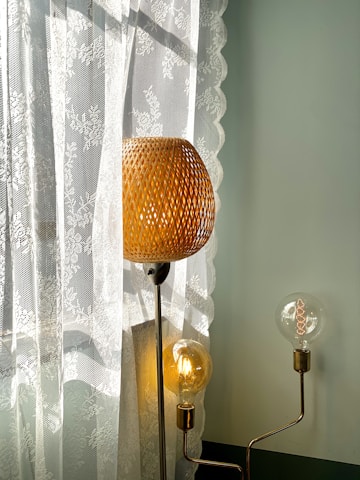 A woven lampshade emits a warm glow, standing beside a sheer lace curtain that features floral patterns. Additional exposed light bulbs add a modern touch with their visible filaments, while soft daylight filters through the lace, creating gentle shadows.