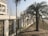 A grand white mansion with ornate balconies and columns is situated behind a decorative wall featuring intricate wrought iron designs. A palm tree is planted in the foreground, next to a sidewalk that runs along a grassy area.