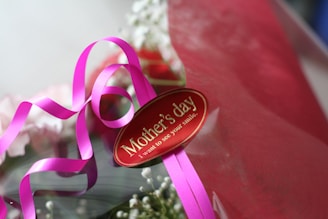 A bouquet wrapped in red paper adorned with a bright pink ribbon and a tag that reads 'Mother's day. I want to see your smile.' The bouquet includes white flowers and hints of other colors, creating a festive and celebratory presentation.