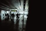 Silhouettes of people walking with luggage in a modern, dimly lit train station or airport terminal. The reflective floor and large windows create a dramatic contrast against the bright exterior light shining through. Overhead signs display travel information.