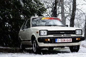 A vintage white Toyota car is parked outdoors on a snowy day, surrounded by snow-covered trees and falling snowflakes. The car has a colorful decal on its windshield, and its headlights are distinctive and rectangular. The license plate reads '1-SUN-648'. The setting is wintry and serene, with a background of leafless trees and a lush evergreen tree partially covering the car.