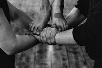 Four arms join together, with hands gripping each other's wrists, forming a strong bond or union. The image is in black and white, highlighting the contrasts and textures of the skin and clothing.