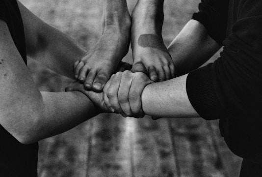 Four arms join together, with hands gripping each other's wrists, forming a strong bond or union. The image is in black and white, highlighting the contrasts and textures of the skin and clothing.