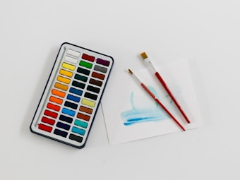 A set of watercolor paints with a variety of colors arranged in a grid is seen alongside two red-handled paintbrushes resting on a piece of paper with a light blue brushstroke.