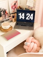 A cozy workspace features a laptop displaying a digital clock reading 14:24. The desk is adorned with makeup products, brushes, and a cup of coffee. A stylish pink and gold notebook lies next to the laptop. Floral art prints and motivational quotes decorate the background, along with a plush pink knot pillow on a white chair.