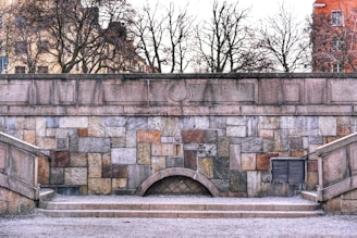 A stone wall composed of multicolored rectangular blocks with an arch design near the bottom center. The wall is framed by two stone staircases on either side, covered in light snow or frost. Bare trees stand behind the wall, with a building partially visible on the right side.