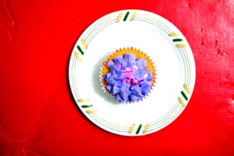 A cupcake with purple frosting topped with small decorative pink beads sits on a decorated white plate. The plate has green and yellow accents and is placed on a bright red textured surface.